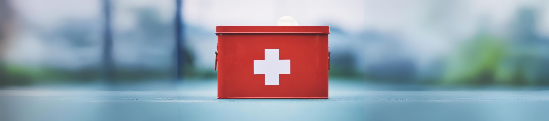 A red first aid kit with a white cross symbol sits on a flat surface, accompanied by effervescent cough tablets, with a blurred outdoor background.