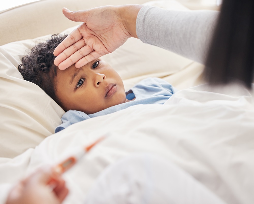 A young child lies in bed covered with a white blanket, looking unwell. An adult hand checks the childs forehead for a fever, while another holds Prospan cough syrup and a thermometer in the foreground.
