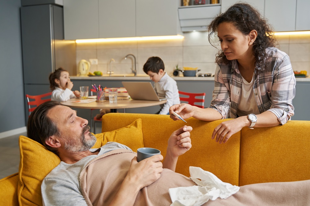 A woman gives prospan to a man lying on a yellow couch with a blanket, while two children eat and do homework at a table in the background in a cozy kitchen.