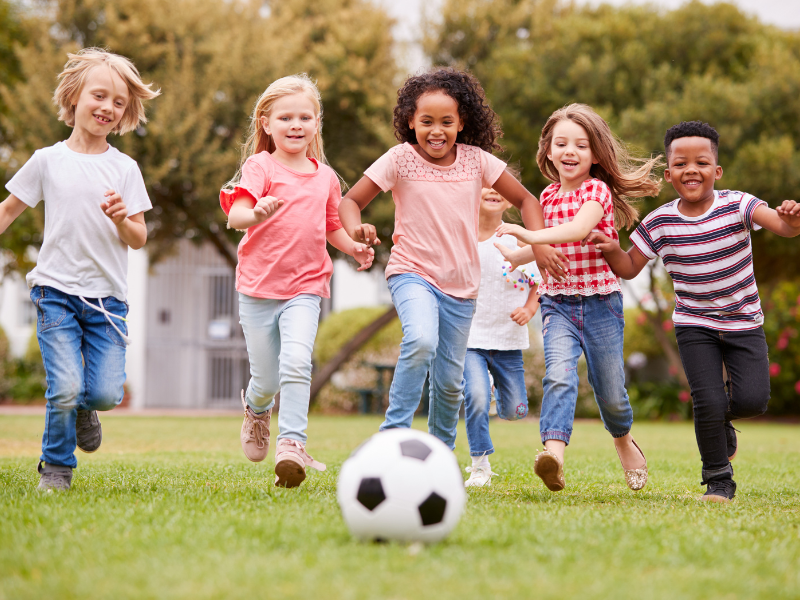 A group of five children run across a grassy field, smiling and chasing a soccer ball on a sunny day. Trees and a building are visible in the background.