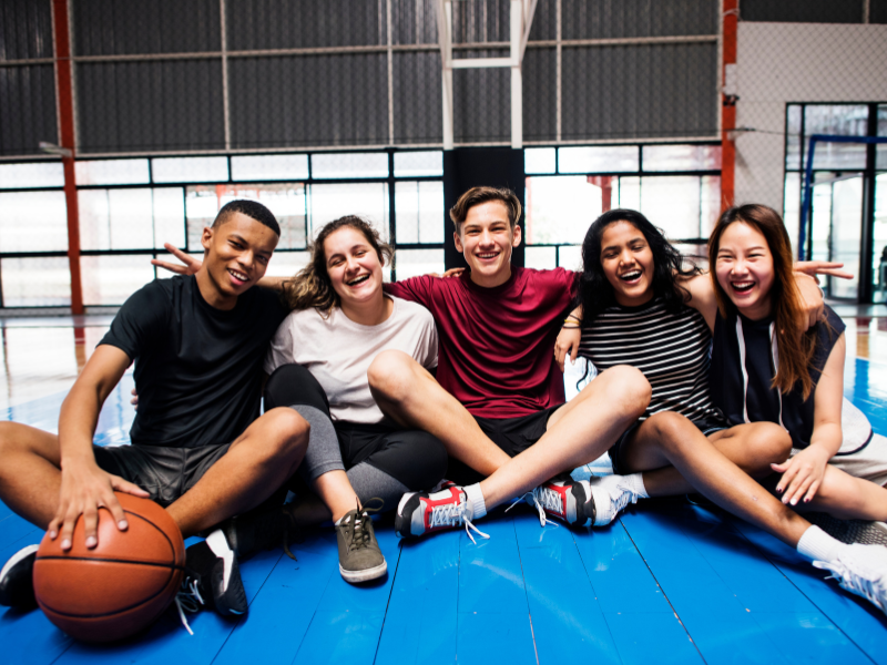 Five teenagers sit together on a basketball court, smiling and laughing with their arms around each other. One holds a basketball, and they appear happy and relaxed in a bright gymnasium.