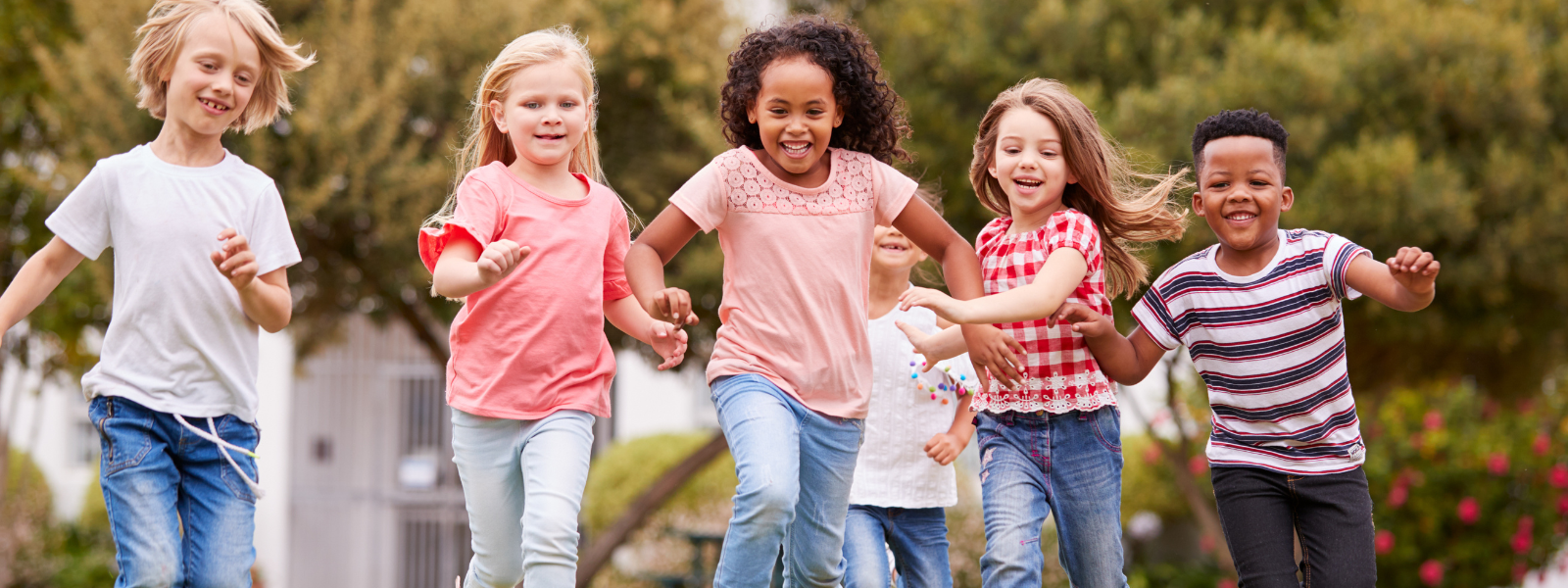 A group of six children, smiling and holding hands, run toward the camera outside in a park, dressed in casual clothes with greenery in the background.
