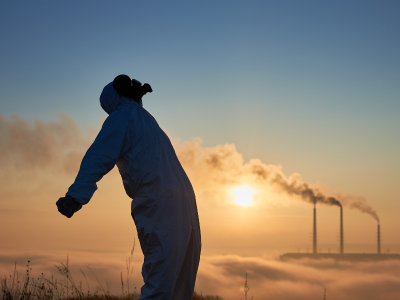 Effects of air pollution on human health. A person wearing a hazmat suit and gas mask stands in a field at sunrise, with smoke billowing from factory chimneys in the background, symbolizing environmental pollution.
