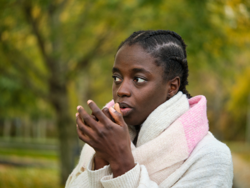 Air quality in South Africa: A person wearing a warm scarf and sweater stands outdoors, holding their hands close to their mouth to keep warm. The background shows blurred green and yellow trees, suggesting a chilly autumn day.