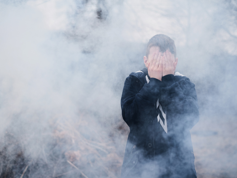 Air quality in South Africa: A person in a dark jacket stands outdoors covering their face with both hands, surrounded by thick, white smoke or mist, with a blurred natural background.